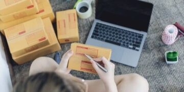 Foto de uma pessoa preenchendo etiquetas de envio em caixas de produtos ao lado de um laptop, representando a criação de uma loja no Instagram Shopping para vender mais em 2024.