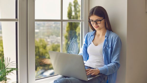 Mulher prestando serviços como social media pelo notebook ao lado da janela