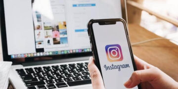 Mão segurando celular com logo do Instagram e notebook ao fundo