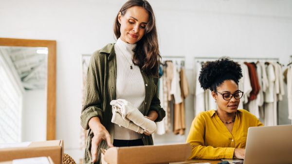 Empreendedora embalando produtos vendidos enquanto profissional da sua equipe escreve posts com frases para loja de roupas