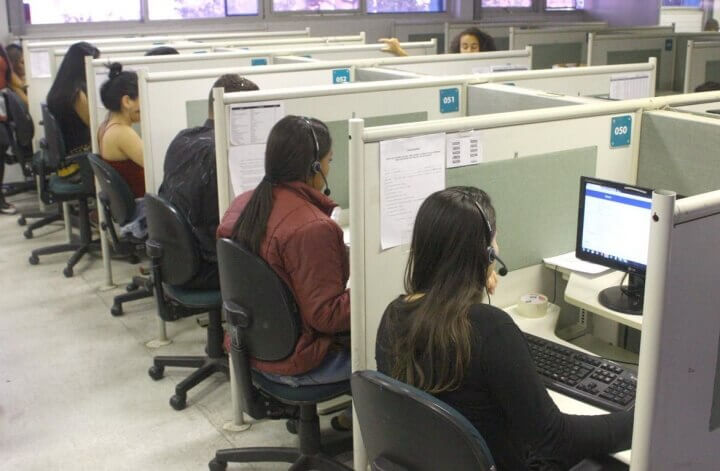 Foto tirada na sede do impulsionegram que mostra diversas pessoas em baias trabalhando no suporte ao cliente